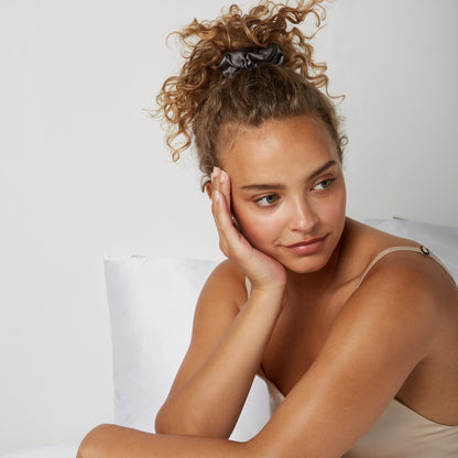 Woman looking happy while sitting comfortably in bed wearing a Luxury Soft Silk  scrunchie from Dore and Rose in the color Gray